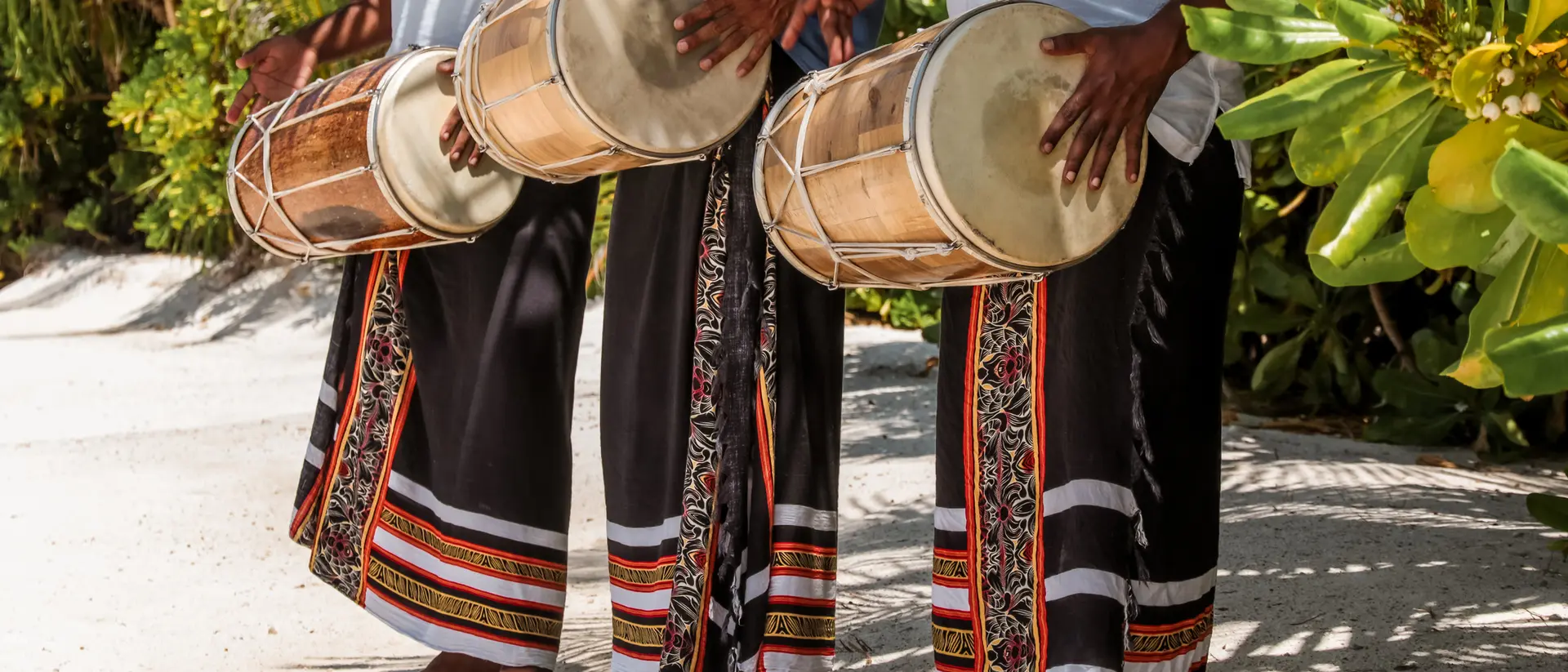 Three musicians in traditional attire playing drums outdoors, surrounded by lush greenery.