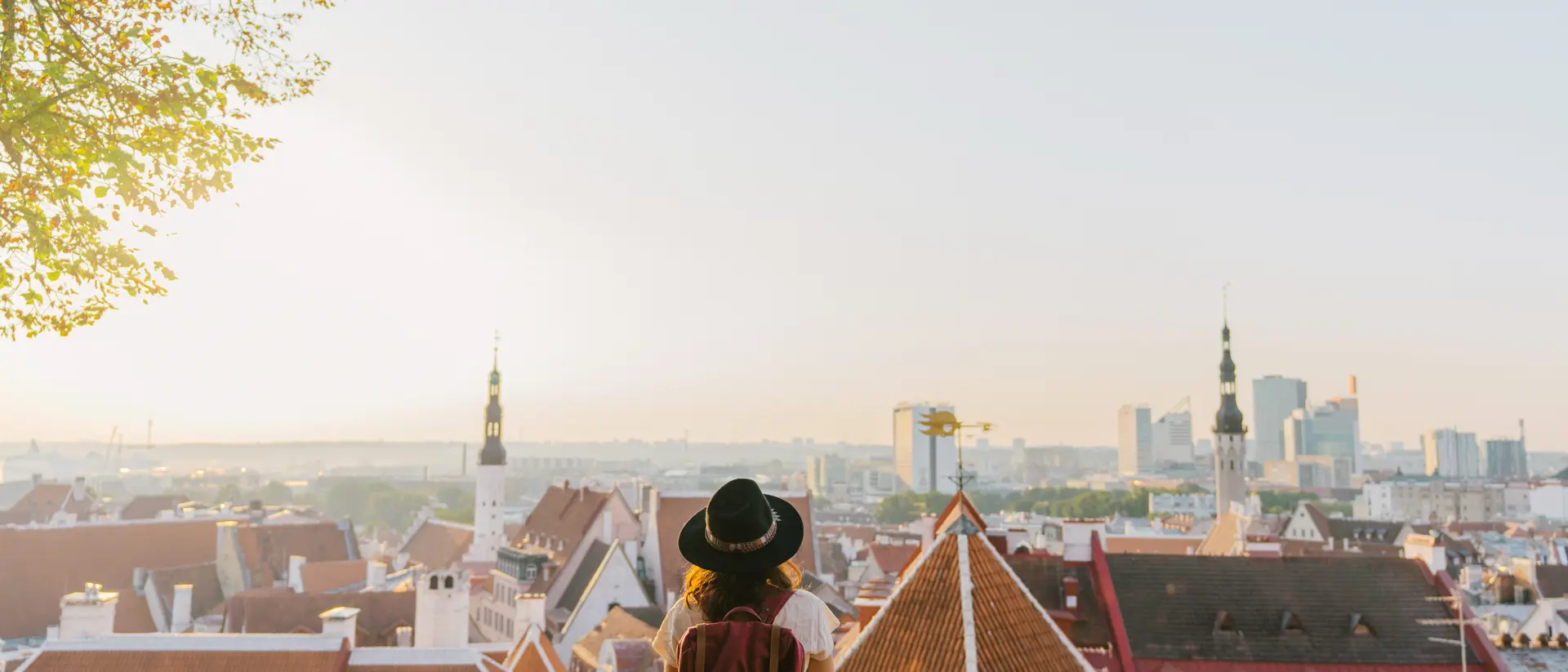 Person in a hat sitting on a ledge, overlooking a cityscape of rooftops and buildings at sunset.