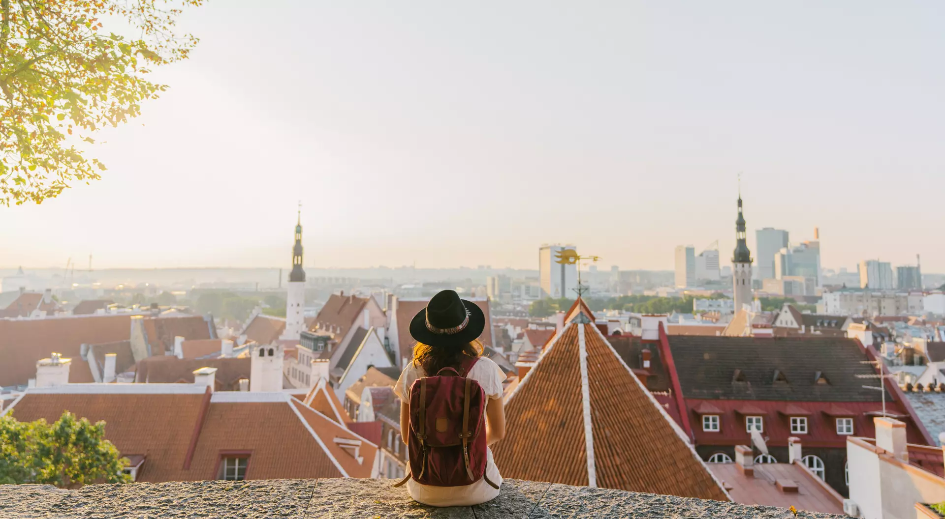 Person in a hat sitting on a ledge, overlooking a cityscape of rooftops and buildings at sunset.