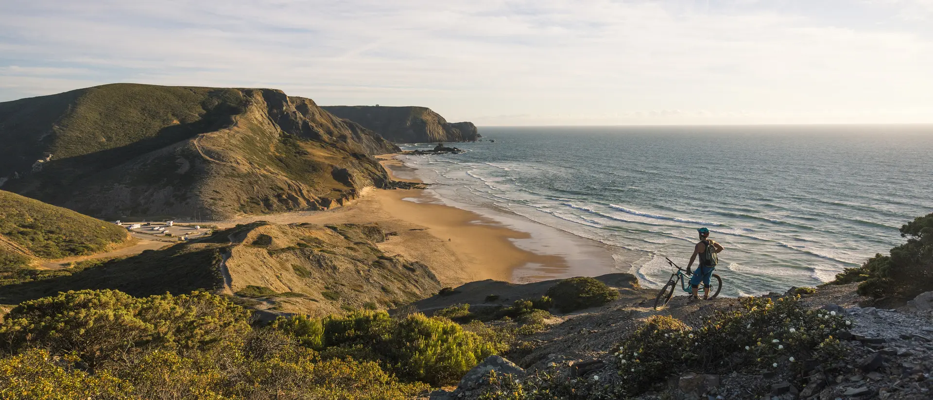 A cyclist stands on a rocky hill overlooking a sandy beach and ocean under a clear sky.