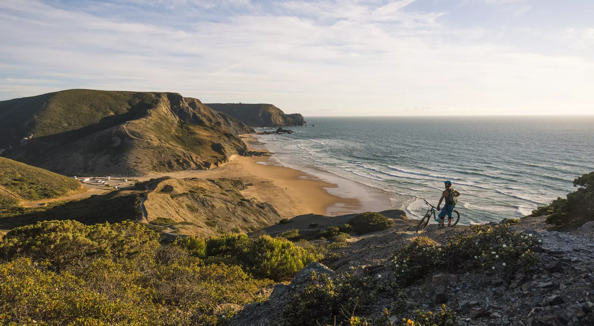 A cyclist stands on a rocky hill overlooking a sandy beach and ocean under a clear sky.