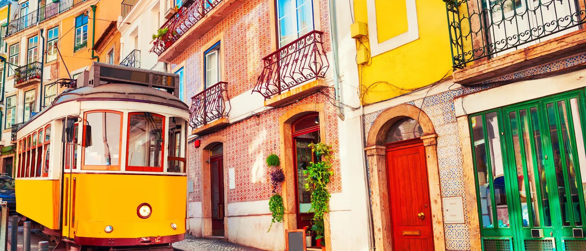 A vintage yellow tram travels along a cobblestone street lined with colorful buildings and balconies in a scenic neighborhood.