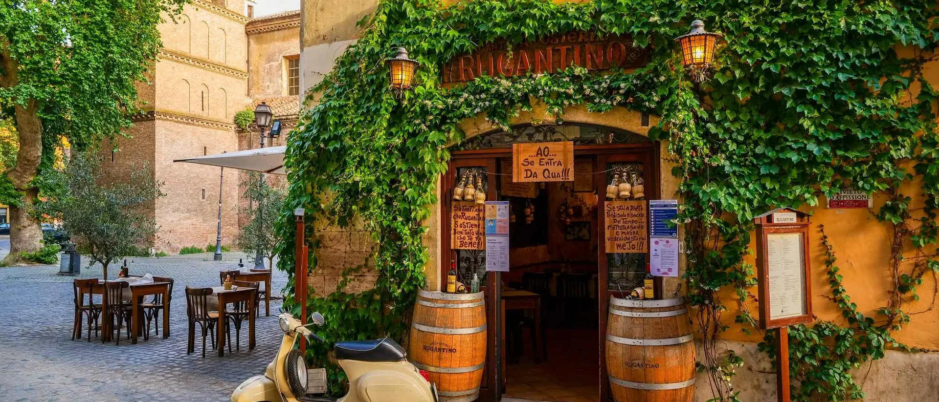Charming outdoor café entrance with greenery, a vintage scooter, and rustic wooden tables in a cobblestone square.