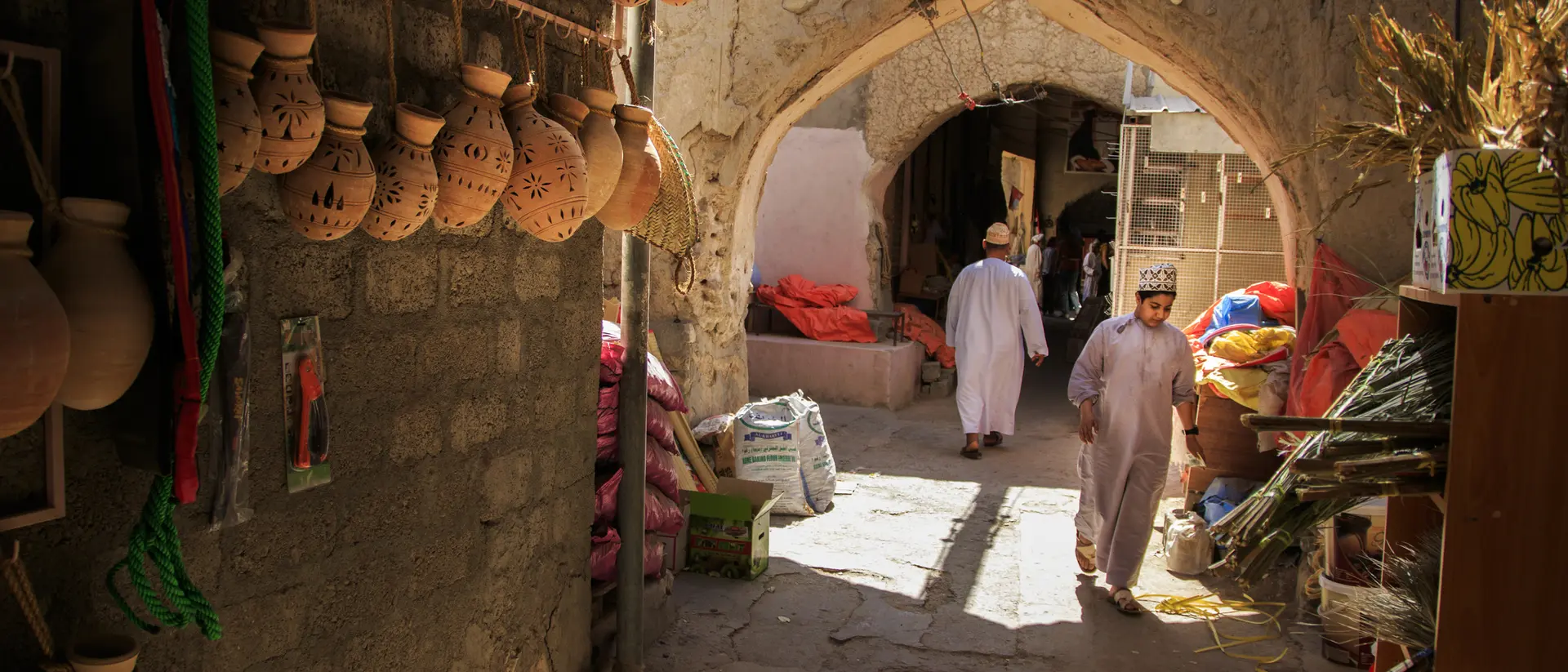 A narrow market alley with clay pots hanging on the wall and people walking through. Brightly colored goods are visible.