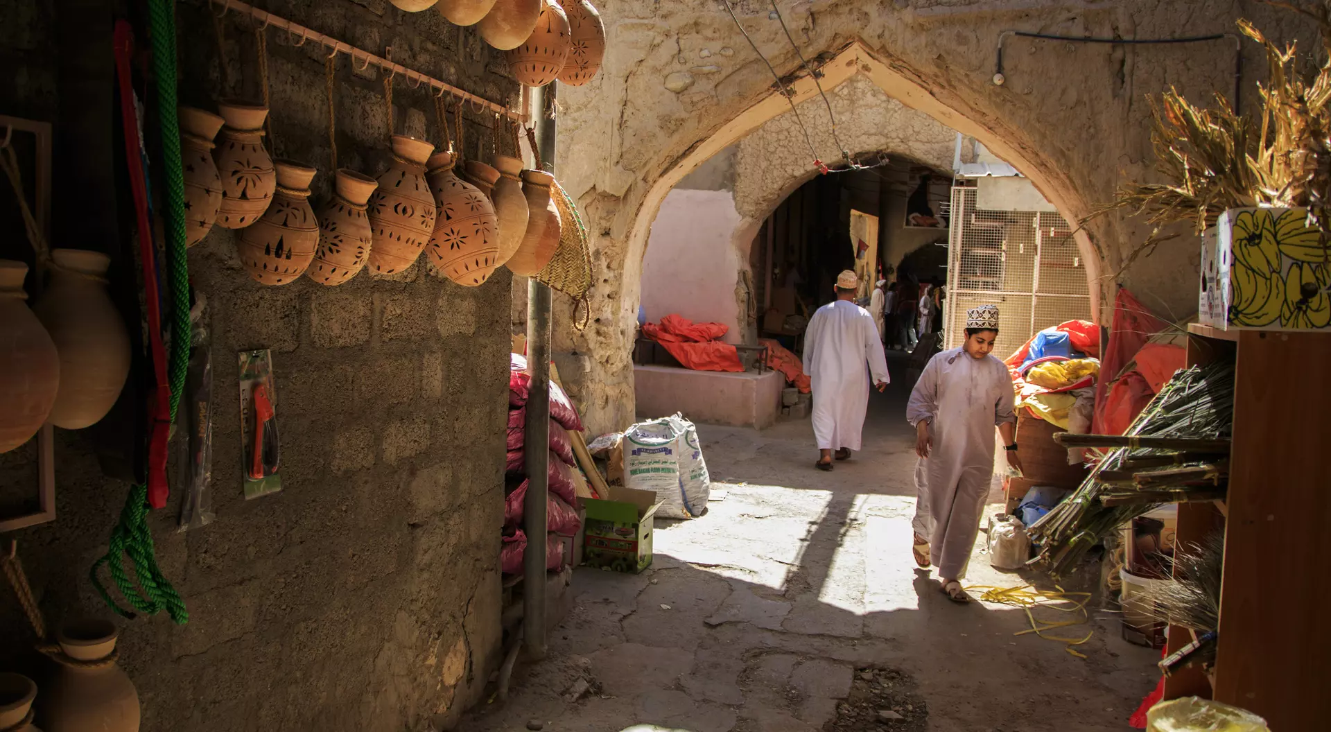 A narrow market alley with clay pots hanging on the wall and people walking through. Brightly colored goods are visible.