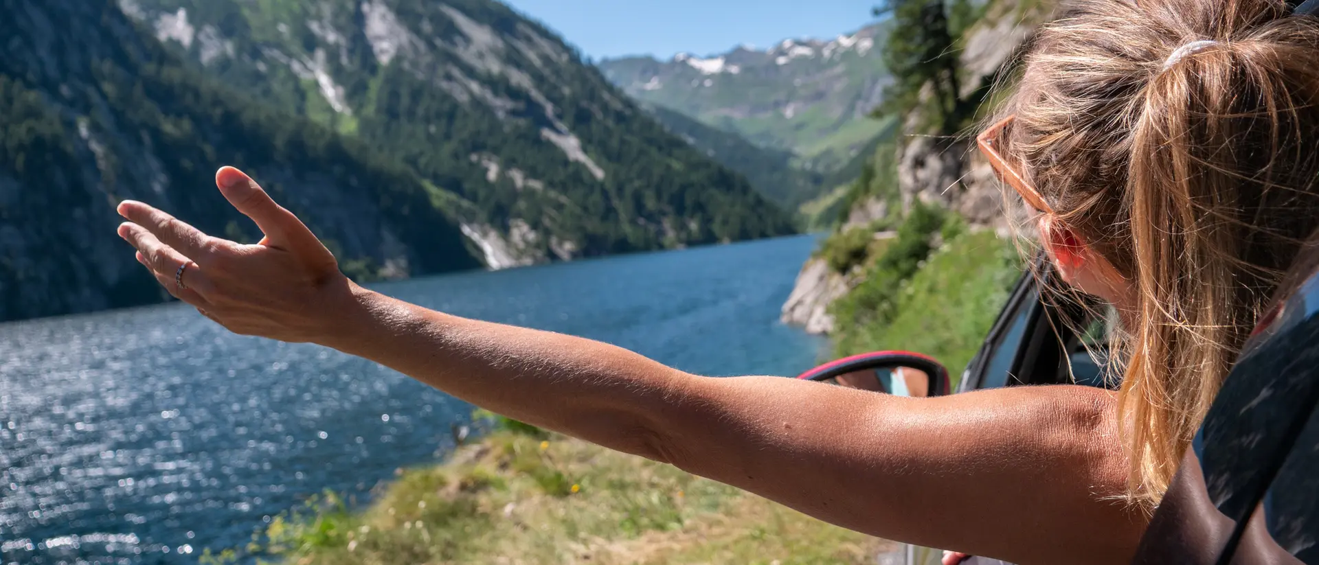 A person leans out of a car window, reaching toward a scenic lake and mountains on a sunny day.