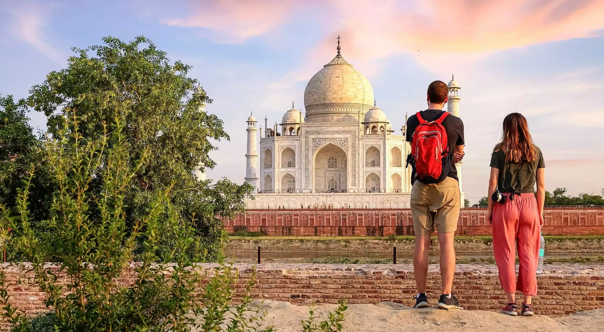 A couple stands on a path, admiring the Taj Mahal against a sunset sky, surrounded by greenery.