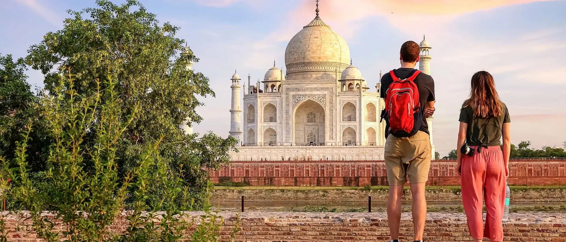 A couple stands on a path, admiring the Taj Mahal against a sunset sky, surrounded by greenery.
