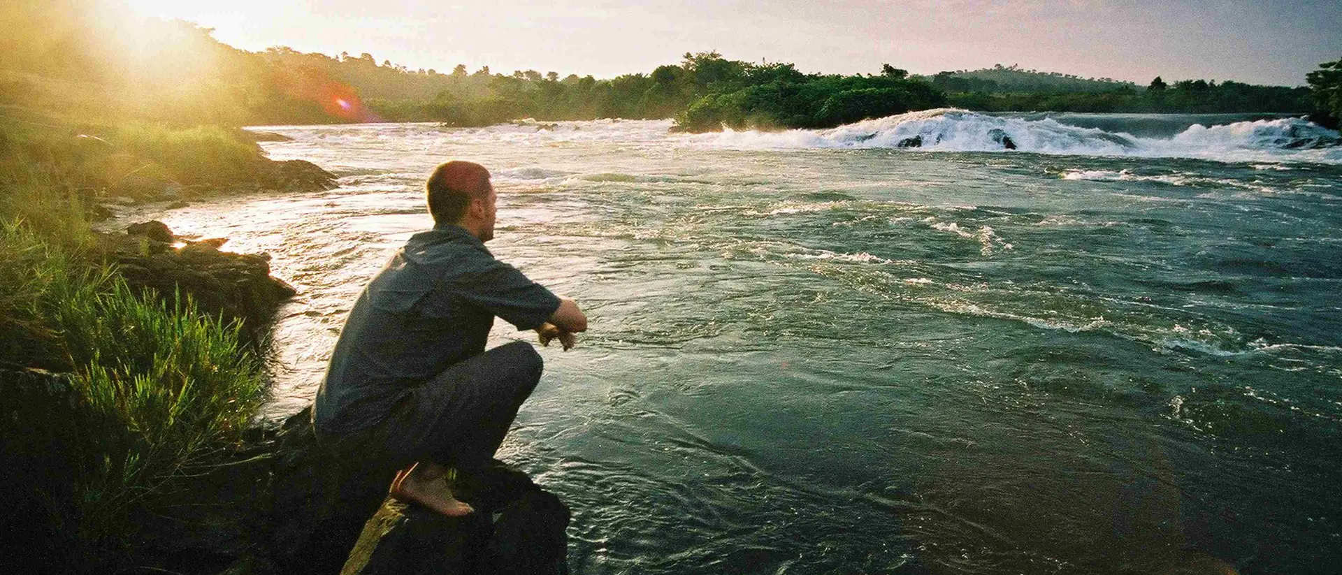 A person sits on a rock by a river, gazing at the water and sunset in a peaceful, natural setting.