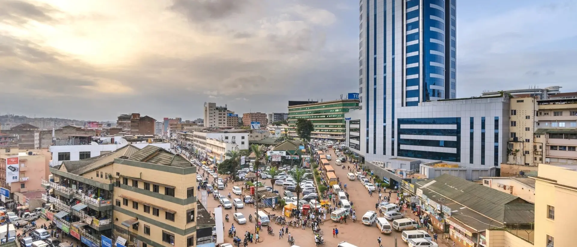 A bustling urban market scene with cars, people, and tall buildings under a cloudy sky during sunset.