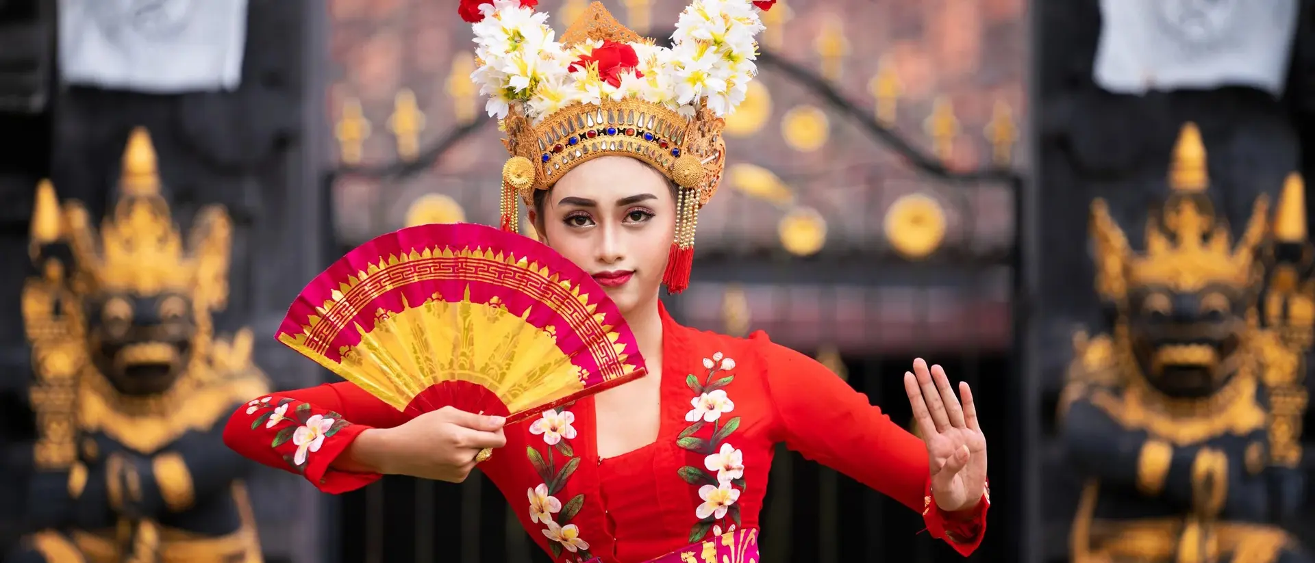 A woman in a vibrant red traditional dress holds a fan while posing in front of ornate temple gates.