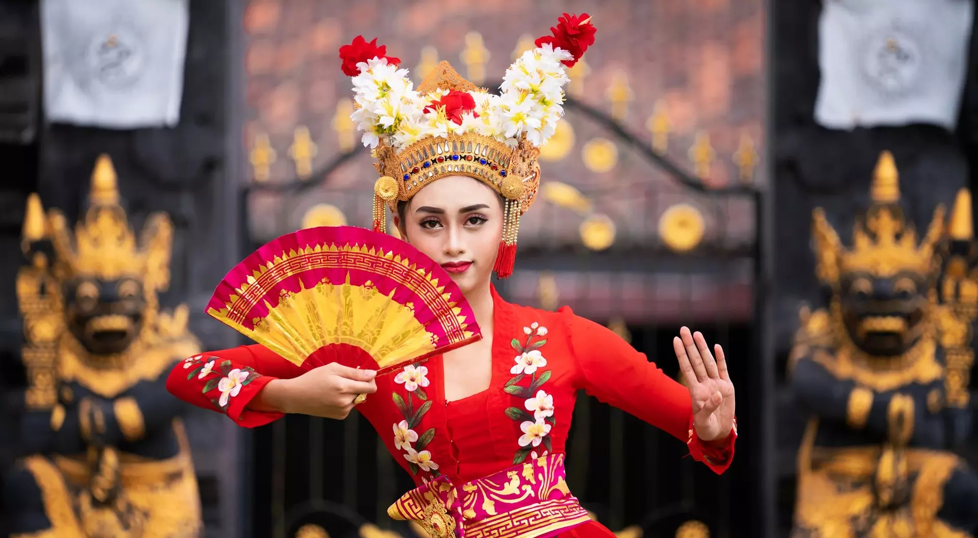 A woman in a vibrant red traditional dress holds a fan while posing in front of ornate temple gates.