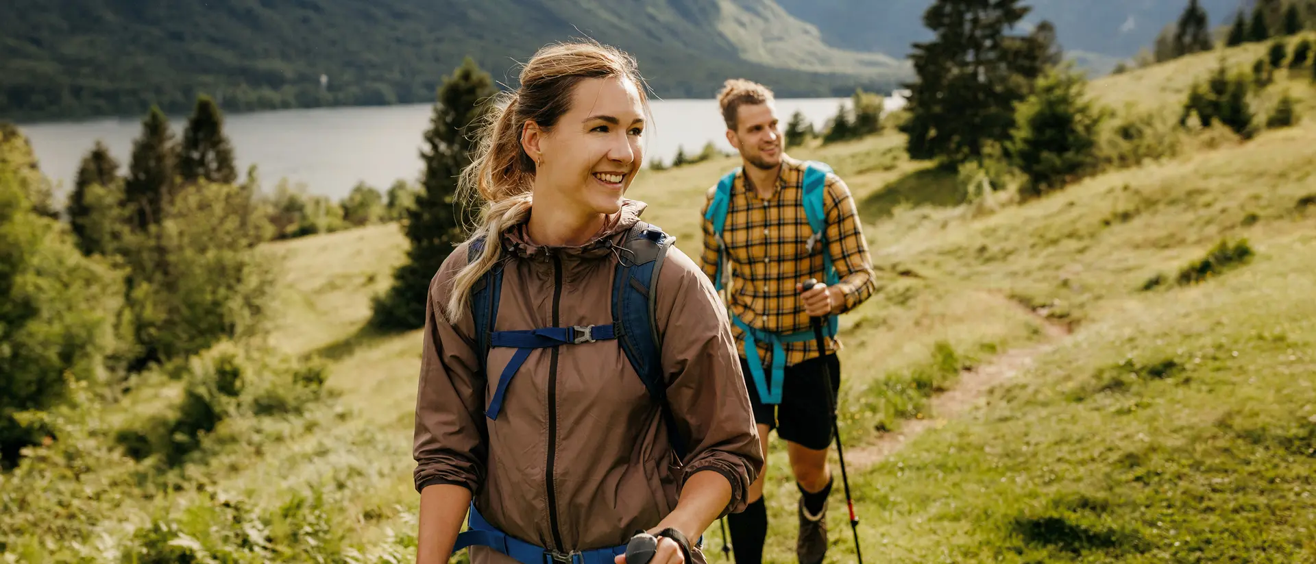 A woman and man hike on a grassy trail with mountains and a lake in the background, enjoying nature.