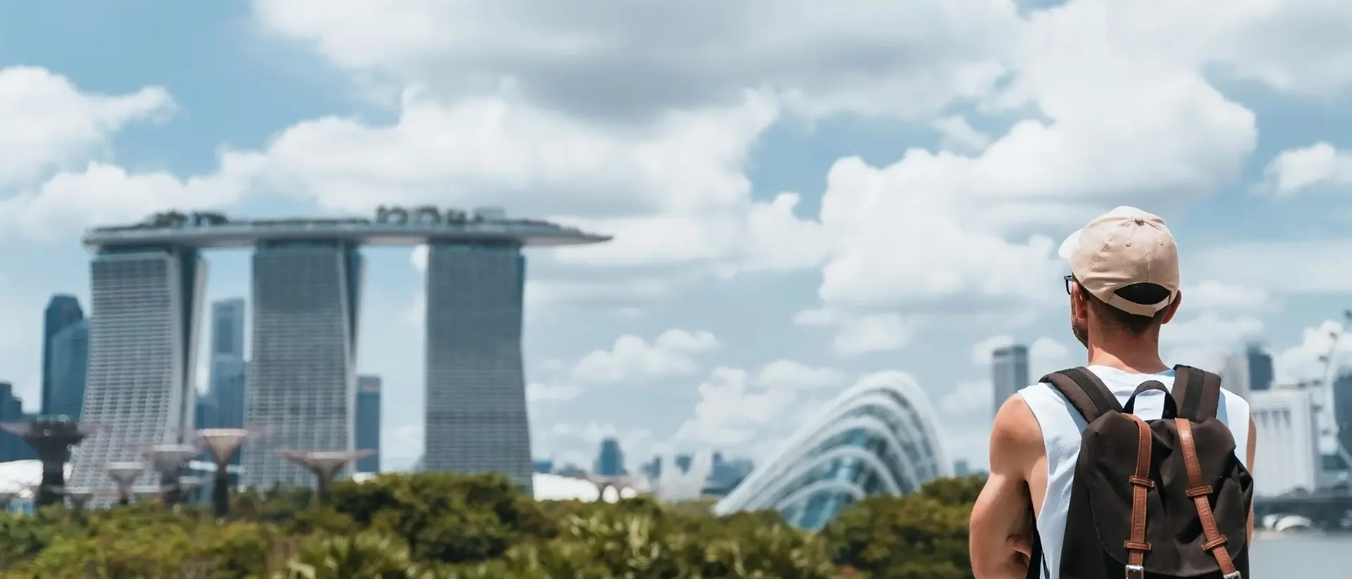A person with a backpack gazes at futuristic buildings under a blue sky with clouds in a lush park setting.