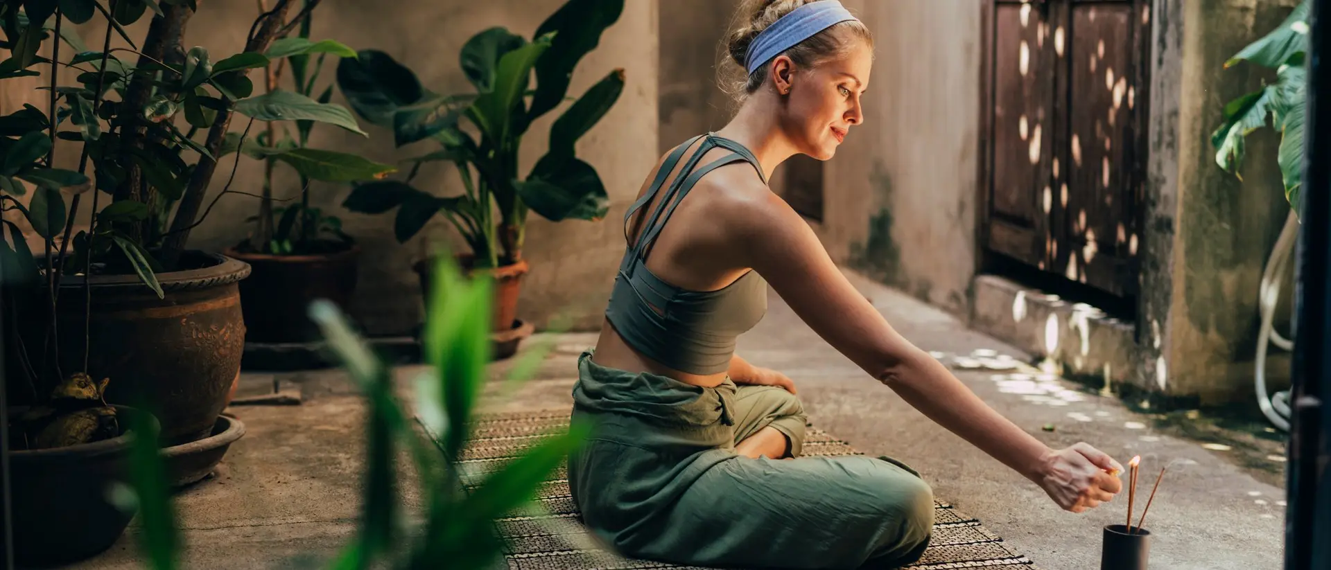 A woman sits cross-legged on the floor in a serene outdoor space, lighting an incense stick surrounded by plants.