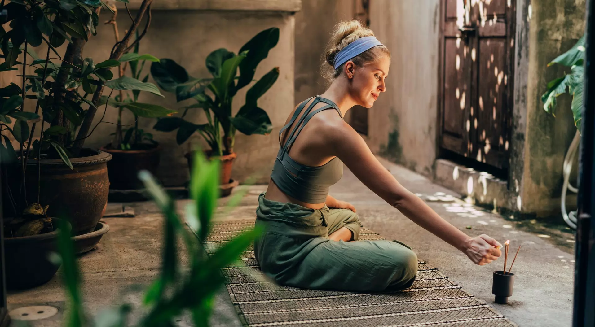 A woman sits cross-legged on the floor in a serene outdoor space, lighting an incense stick surrounded by plants.