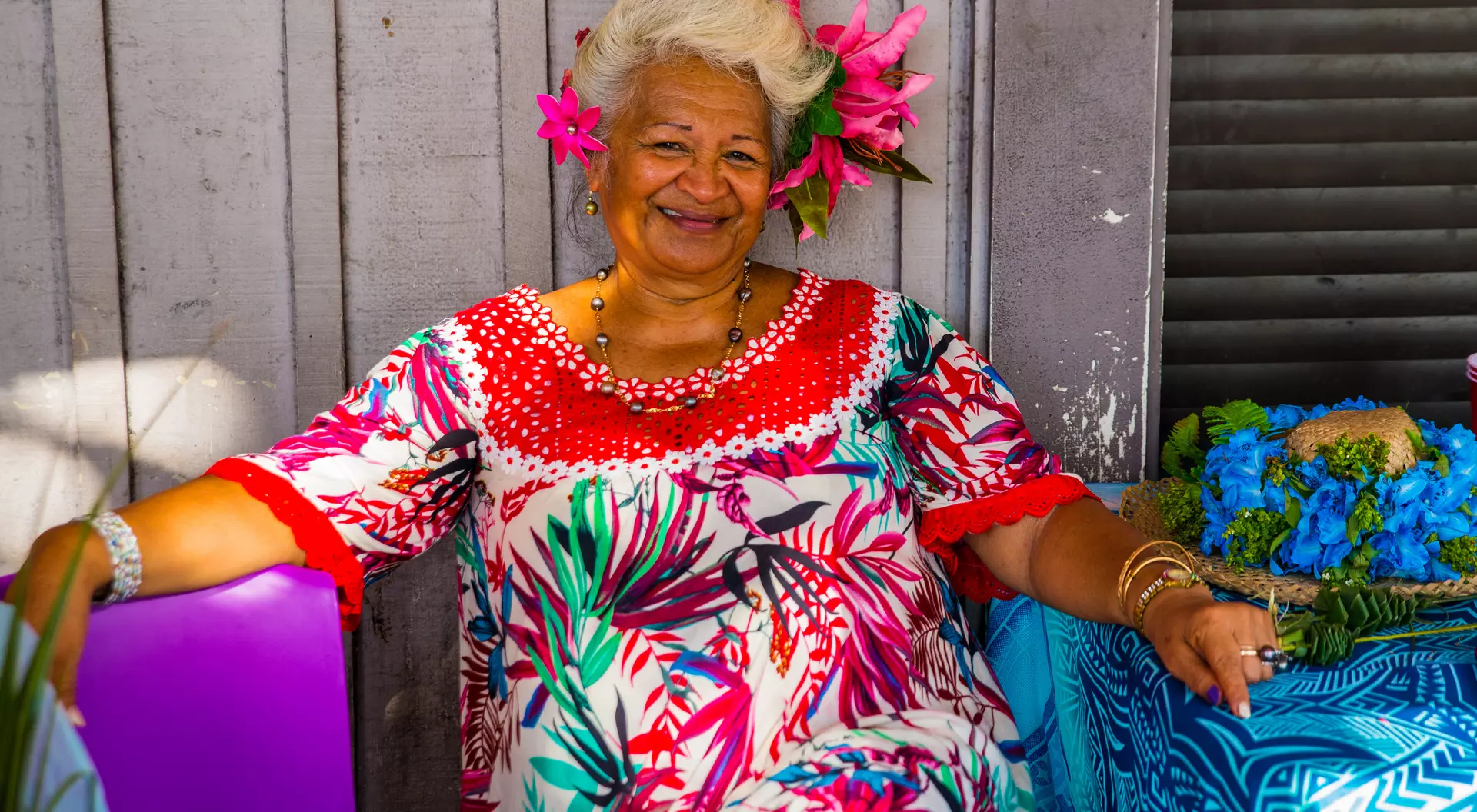 A smiling woman with gray hair and tropical flowers in her hair sits beside a colorful table, wearing a vibrant patterned dress.