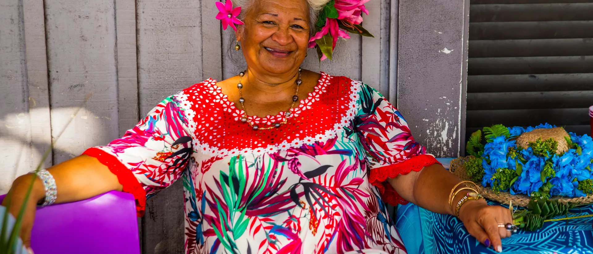 A smiling woman with gray hair and tropical flowers in her hair sits beside a colorful table, wearing a vibrant patterned dress.