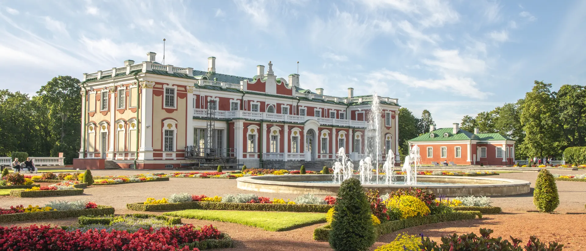 Grand palace with colorful gardens and fountains, under a blue sky, surrounded by blooming flowers.