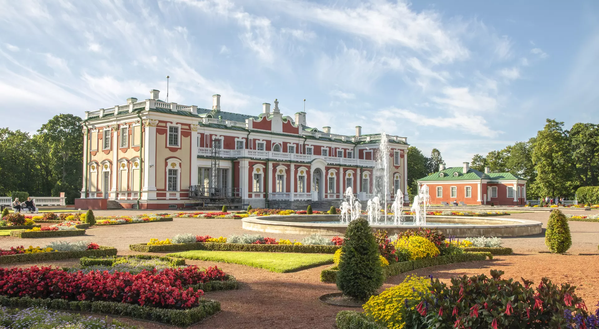 Grand palace with colorful gardens and fountains, under a blue sky, surrounded by blooming flowers.