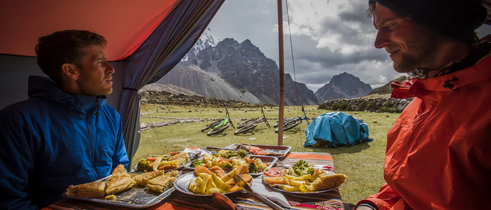Two people sit inside a tent with a table of food, overlooking a scenic mountain landscape.