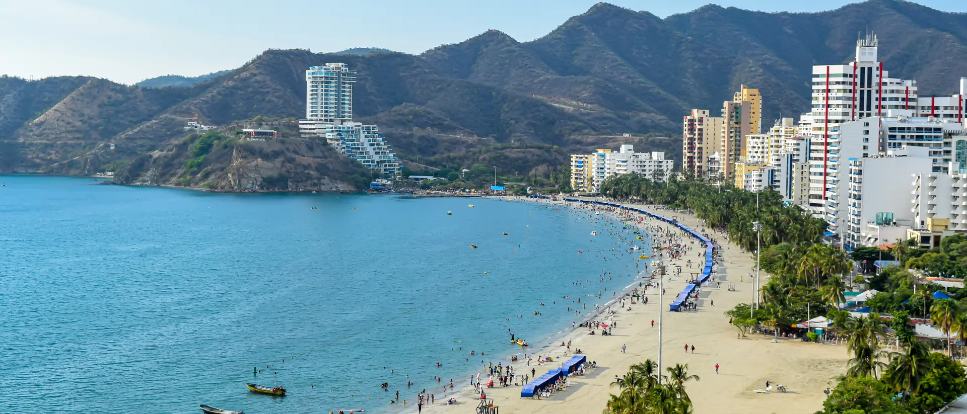 A sunny beach scene with people, boats, and palm trees, surrounded by mountains and buildings along the shoreline.
