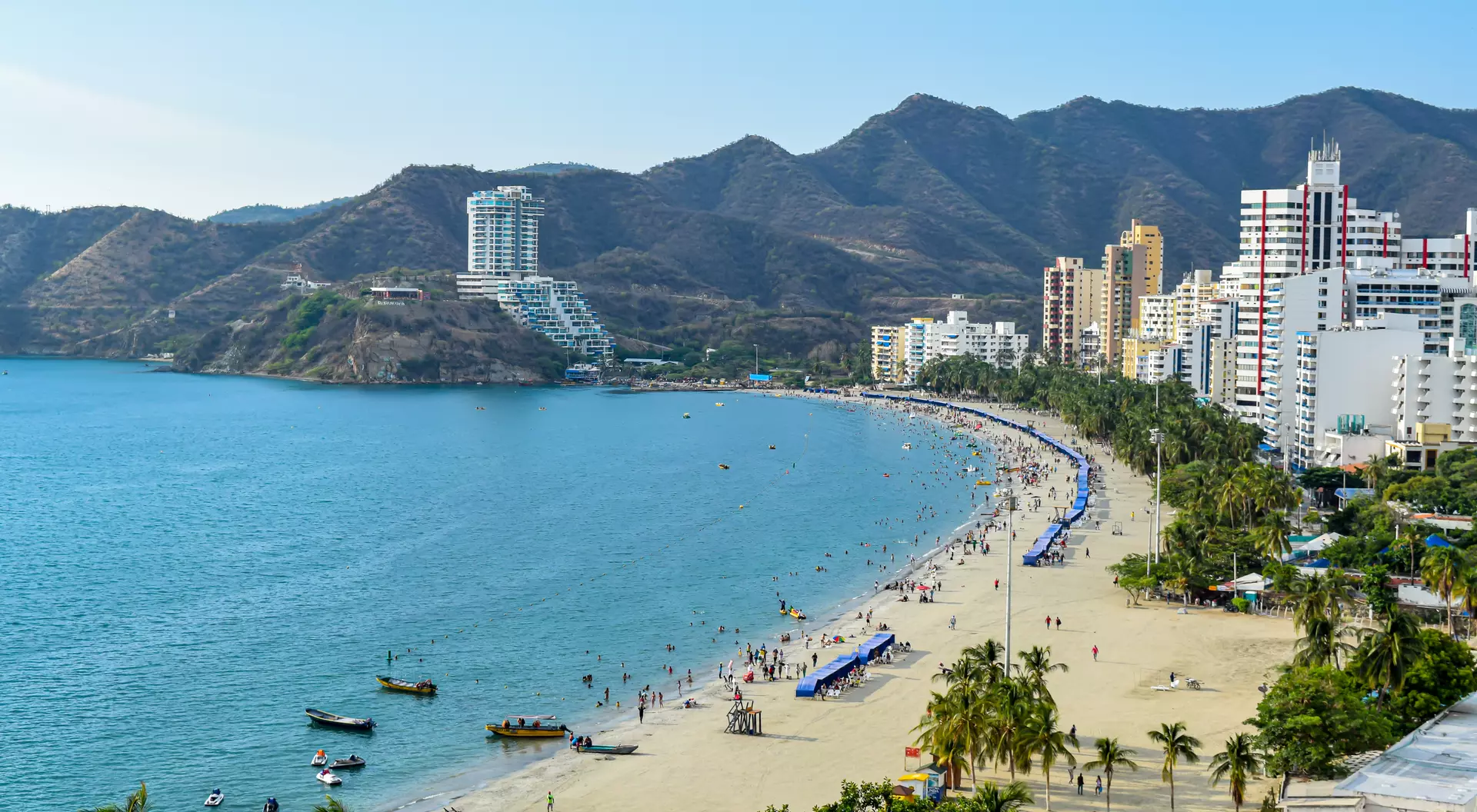 A sunny beach scene with people, boats, and palm trees, surrounded by mountains and buildings along the shoreline.