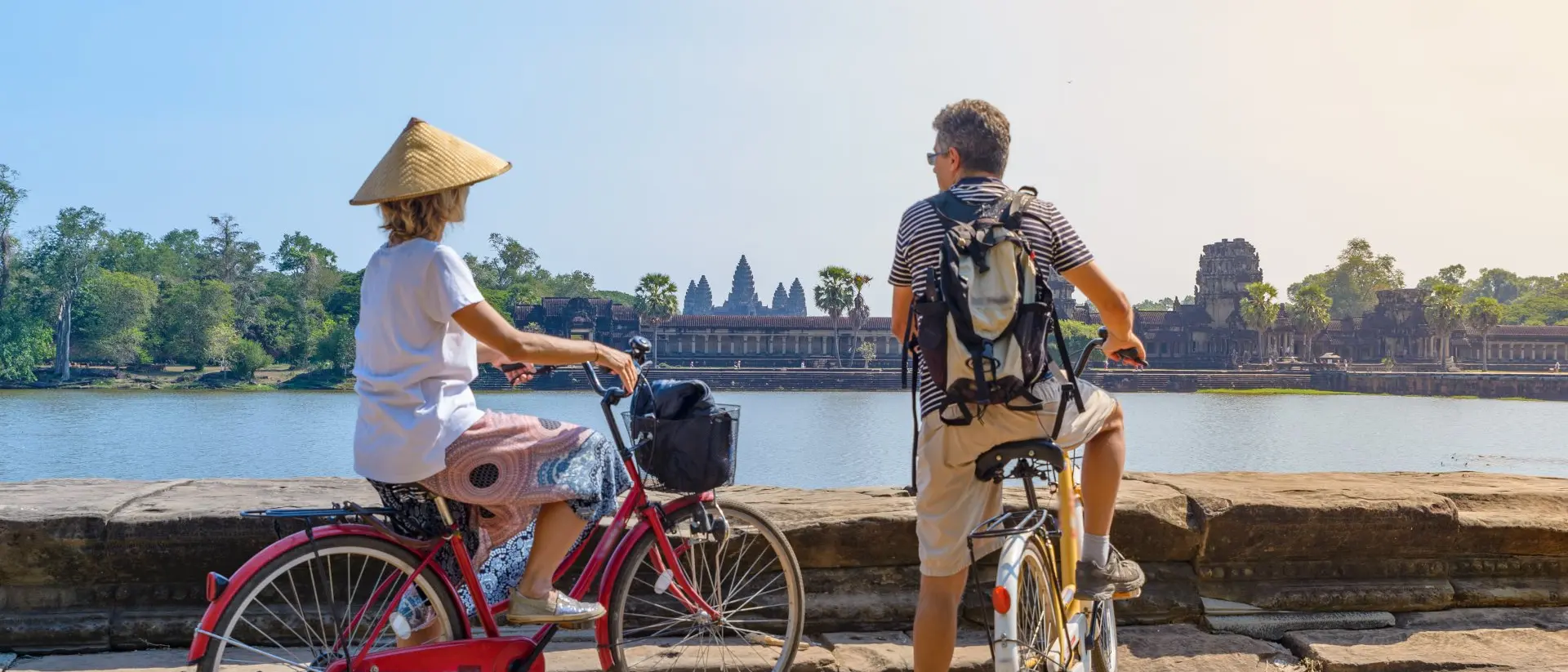 Two cyclists admire a historic temple by a lake in the background, enjoying a sunny day. One wears a traditional hat.