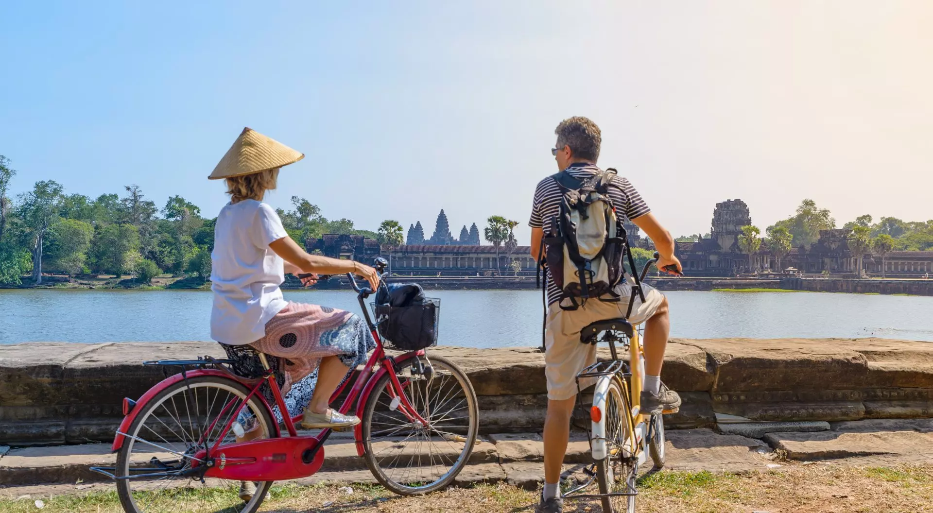 Two cyclists admire a historic temple by a lake in the background, enjoying a sunny day. One wears a traditional hat.