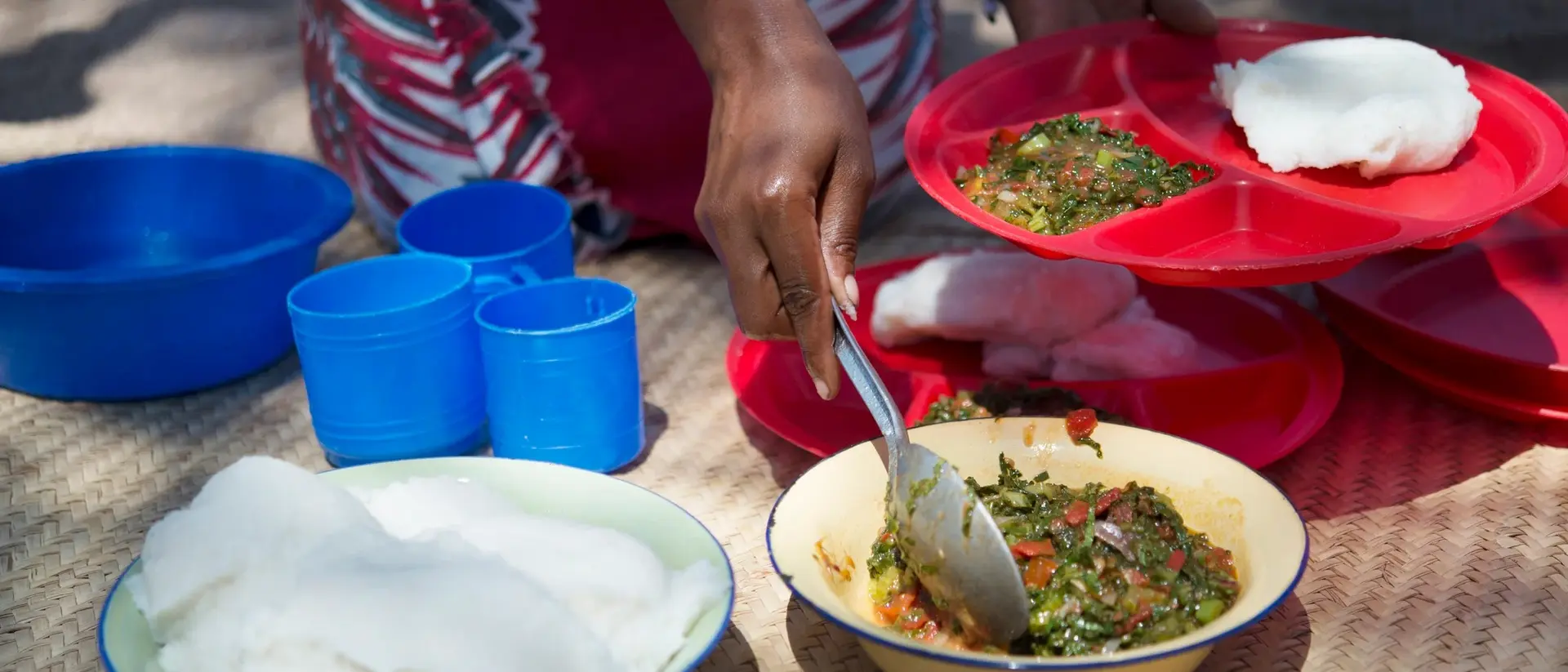 A person serves food from a bowl onto colorful plates, with cups nearby, on a textured surface outdoors.