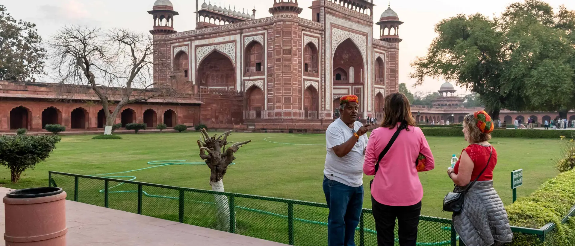 Visitors stand in a landscaped area, talking with an individual near a large red sandstone monument at sunset.