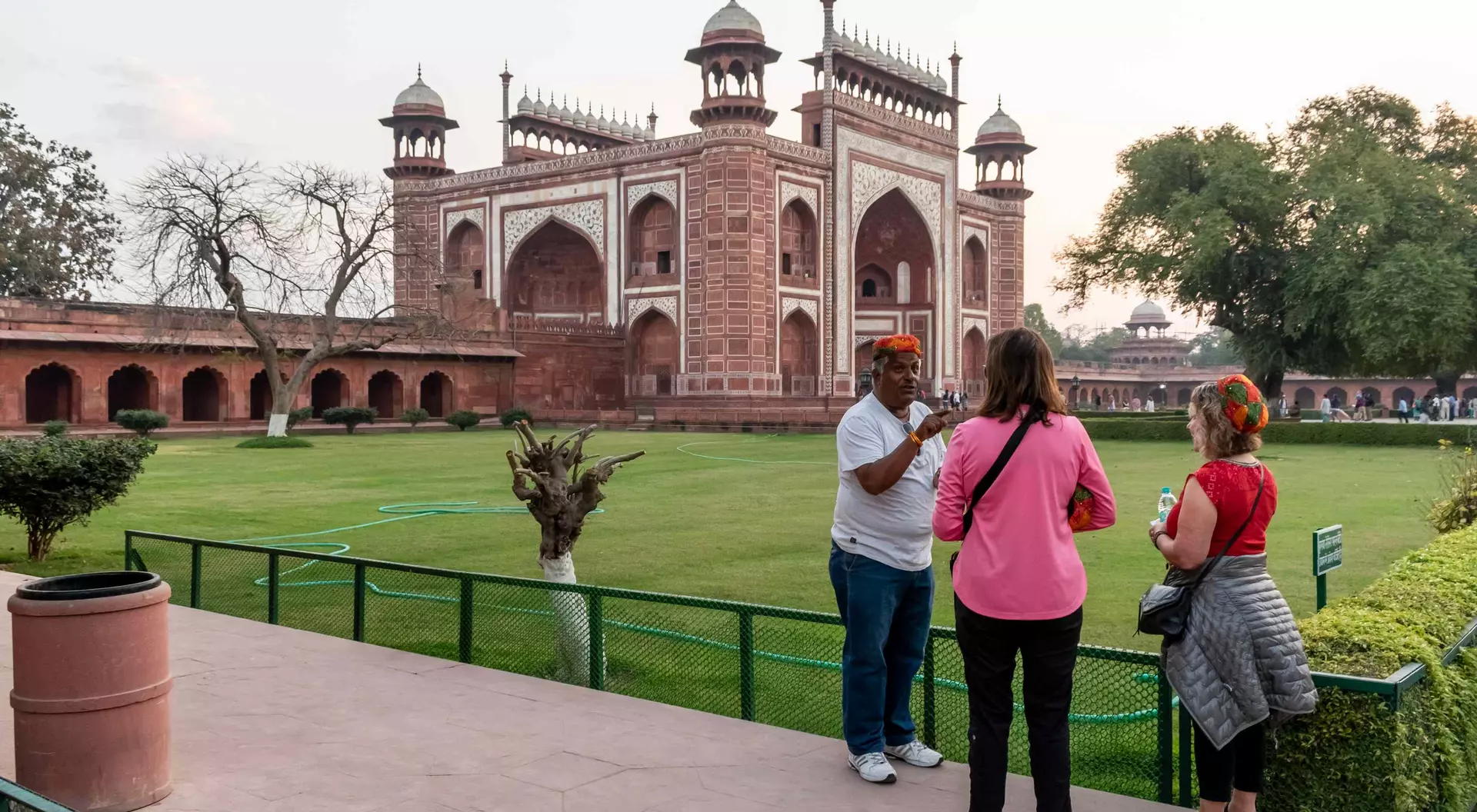 Visitors stand in a landscaped area, talking with an individual near a large red sandstone monument at sunset.