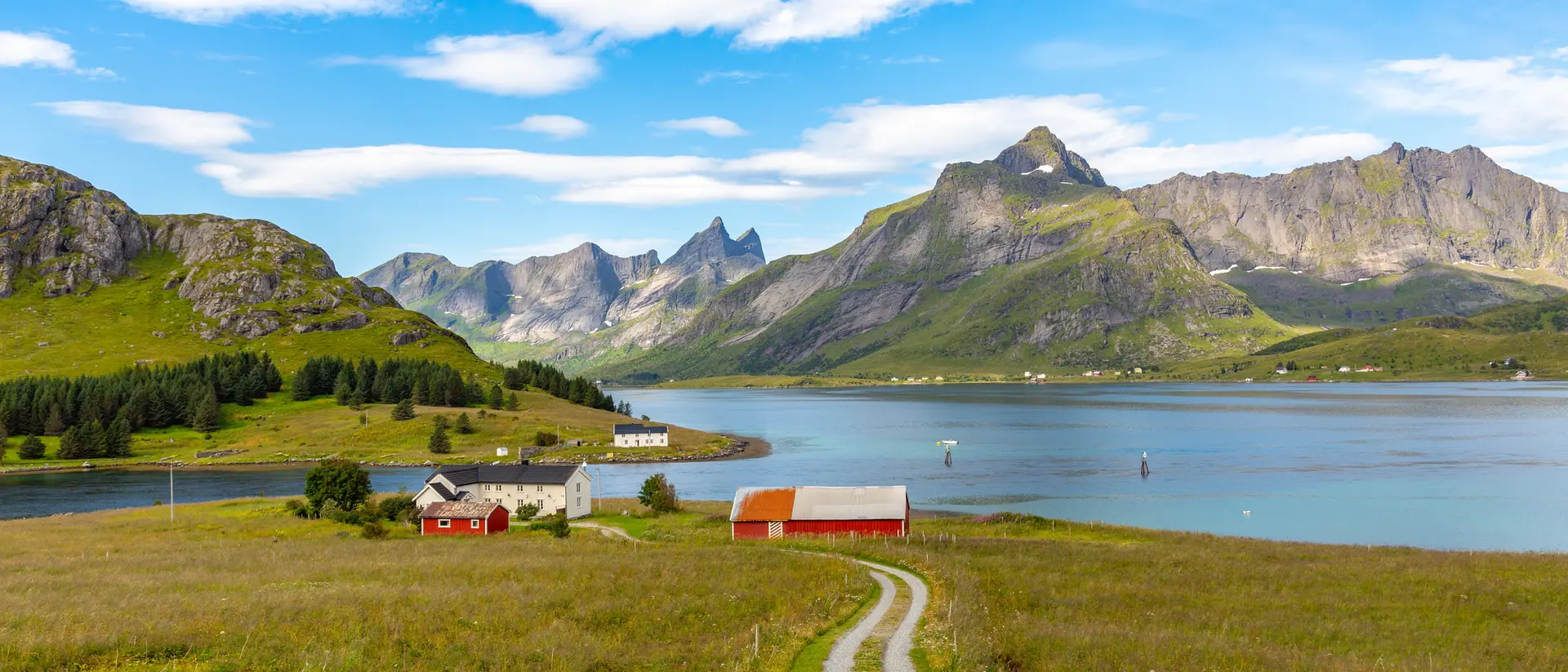 Scenic landscape with mountains, a calm lake, green fields, and red-roofed houses under a blue sky with clouds.