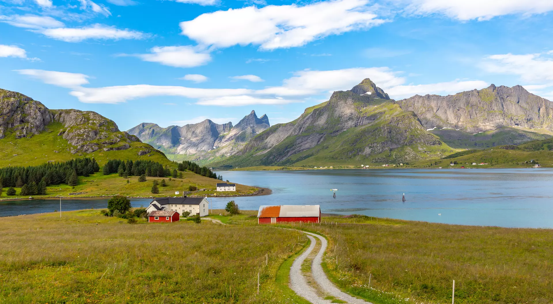 Scenic landscape with mountains, a calm lake, green fields, and red-roofed houses under a blue sky with clouds.