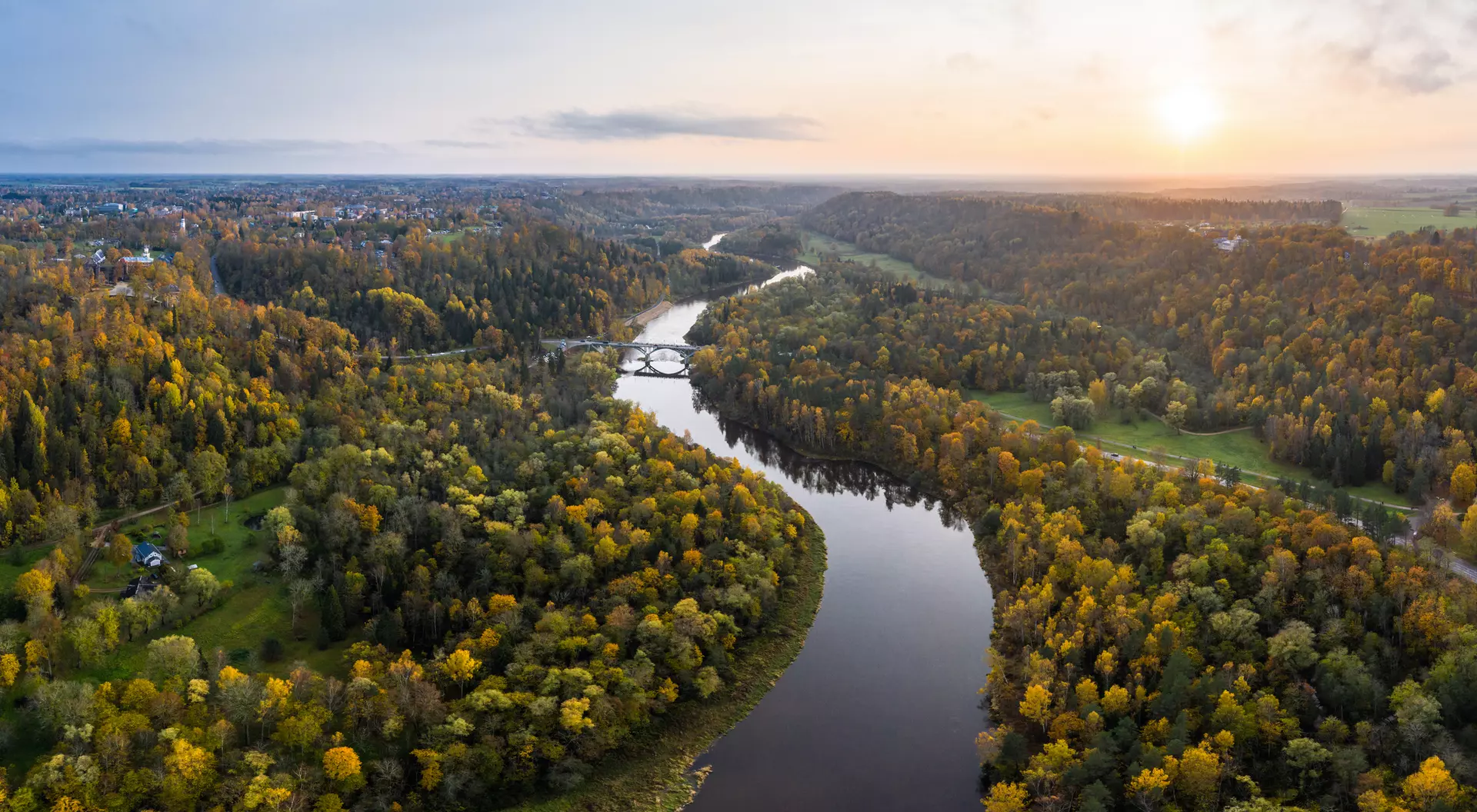 Aerial view of a winding river surrounded by colorful autumn trees and a soft sunset in the background.