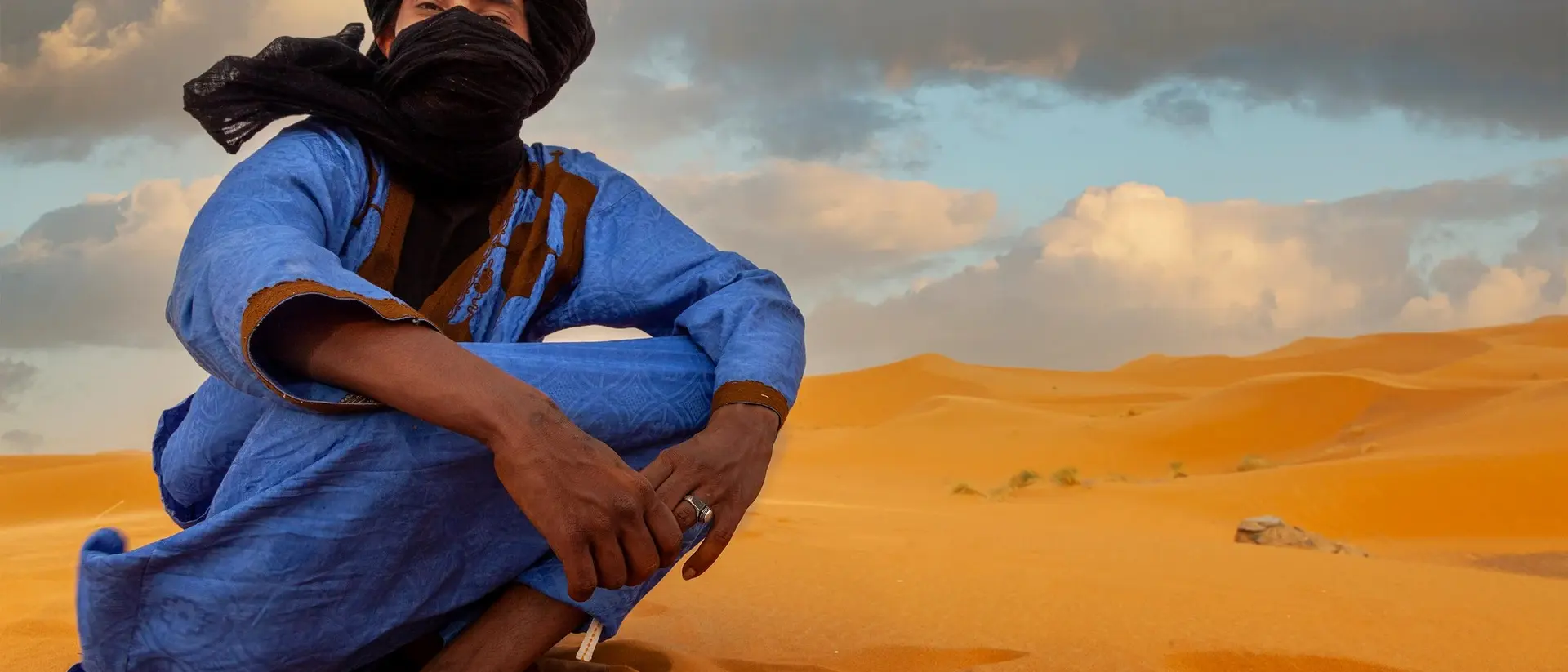 A person in a blue robe and black scarf sits on golden sand dunes under a cloudy sky.