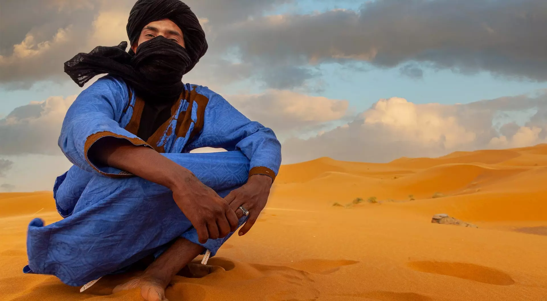A person in a blue robe and black scarf sits on golden sand dunes under a cloudy sky.