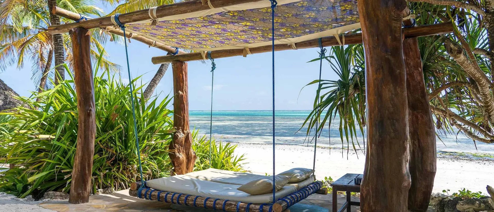 A shaded hammock swing under palm trees on a beach, with clear water and white sand in the background.