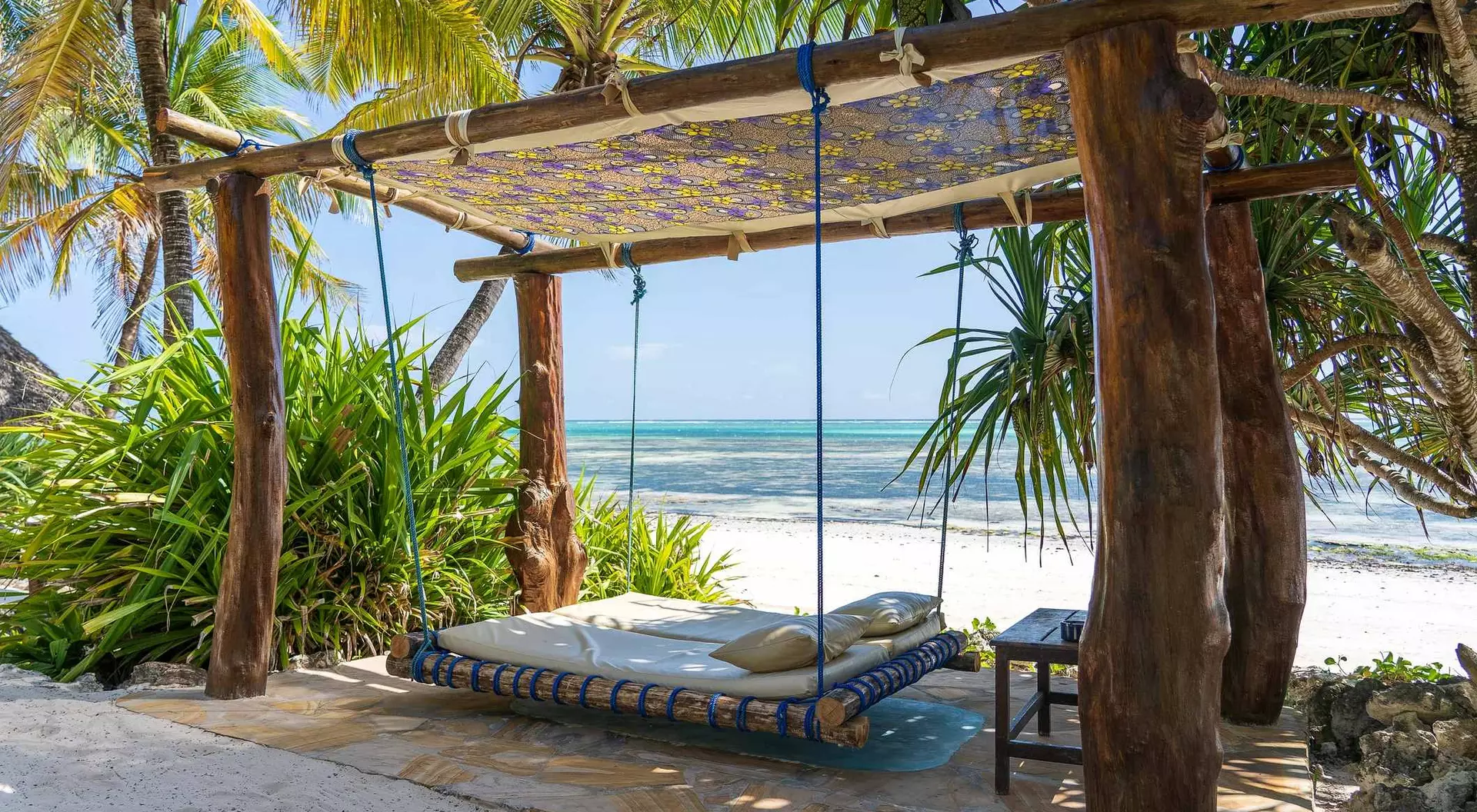 A shaded hammock swing under palm trees on a beach, with clear water and white sand in the background.