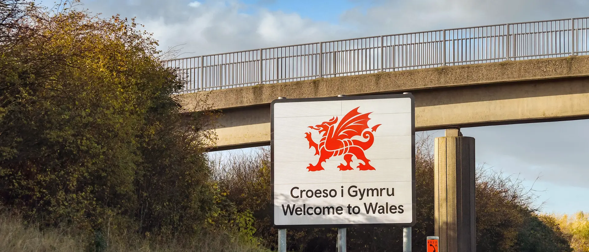 Sign welcoming visitors to Wales with a red dragon emblem, bordered by greenery and a bridge overhead.