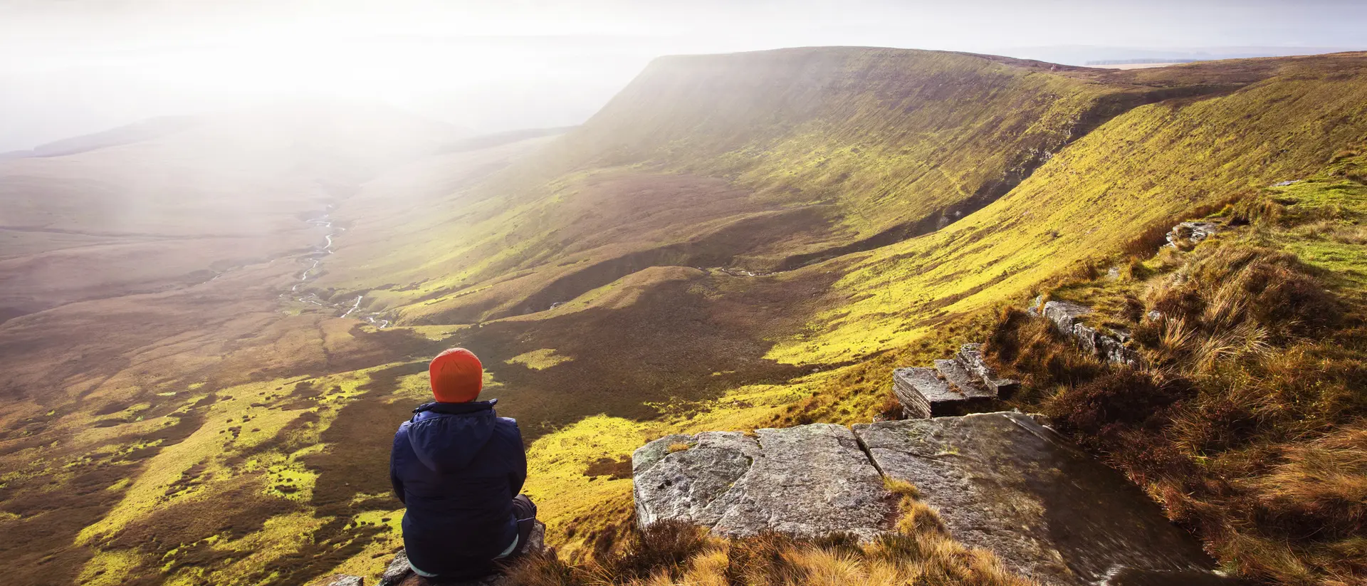 Person sitting on a rocky ledge, gazing at a vast green valley under a cloudy sky.