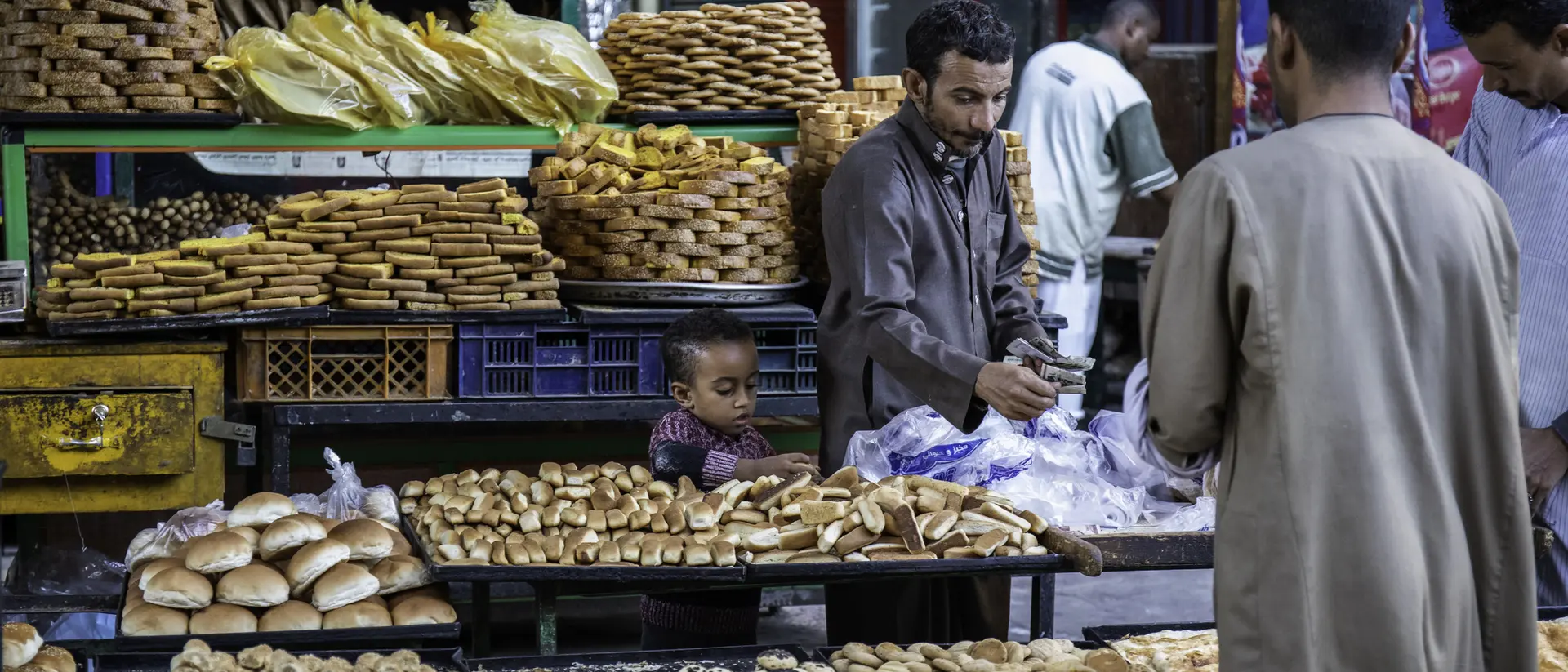 A busy market stall displays various breads and pastries, with a child watching and adults selecting items.