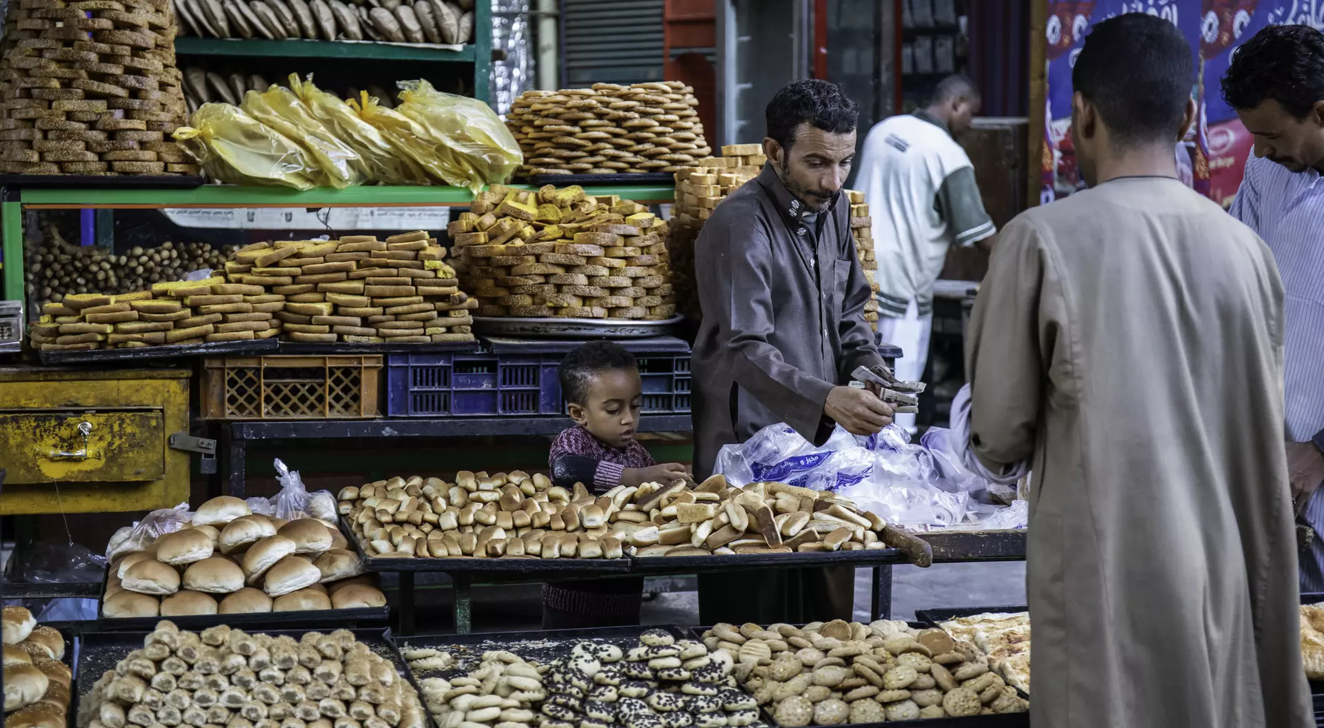 A busy market stall displays various breads and pastries, with a child watching and adults selecting items.
