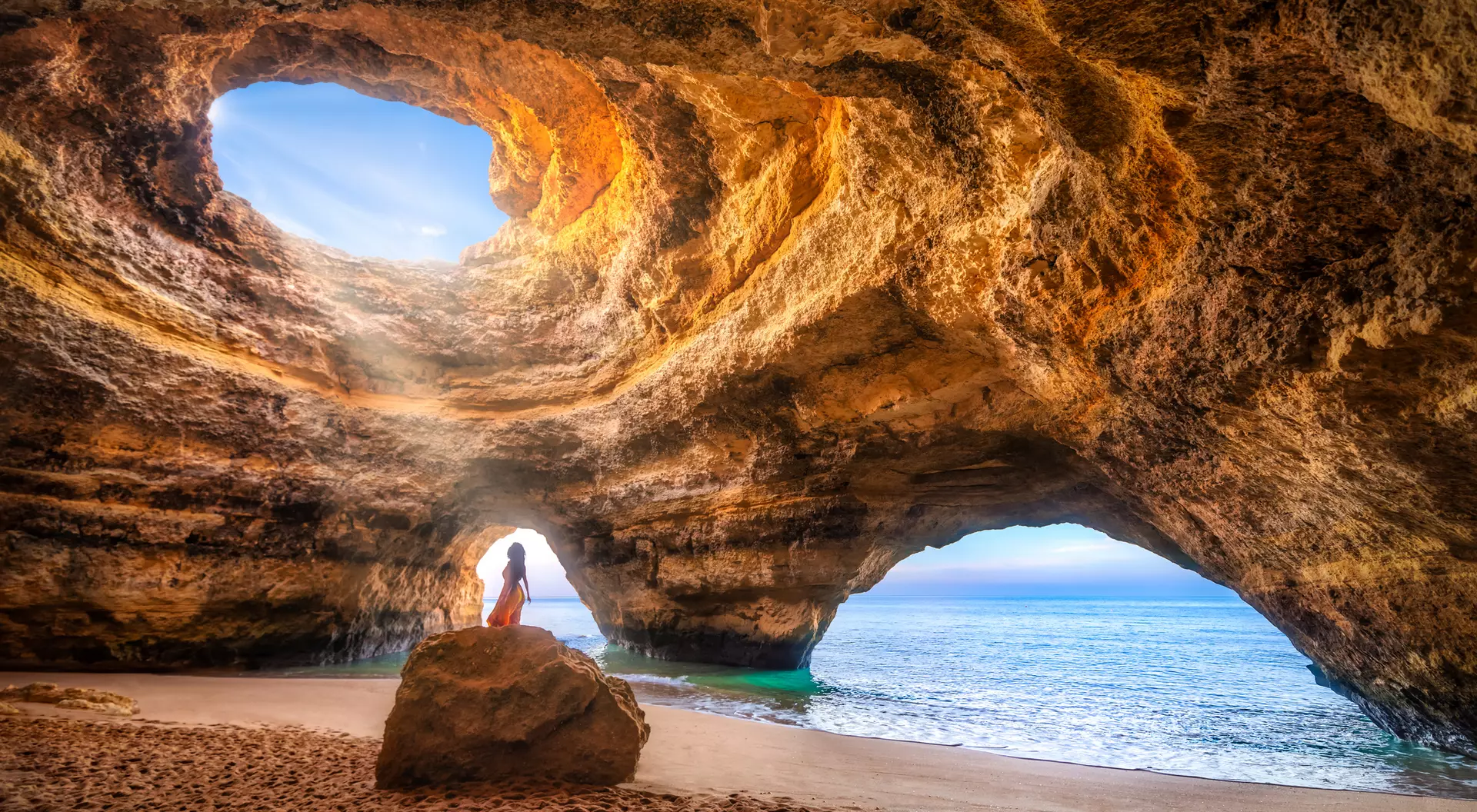 A person stands near a rocky cave opening, looking out at the ocean under a bright sky.