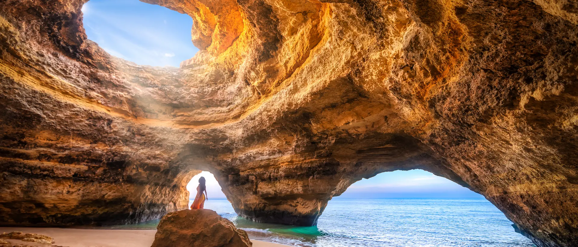 A person stands near a rocky cave opening, looking out at the ocean under a bright sky.