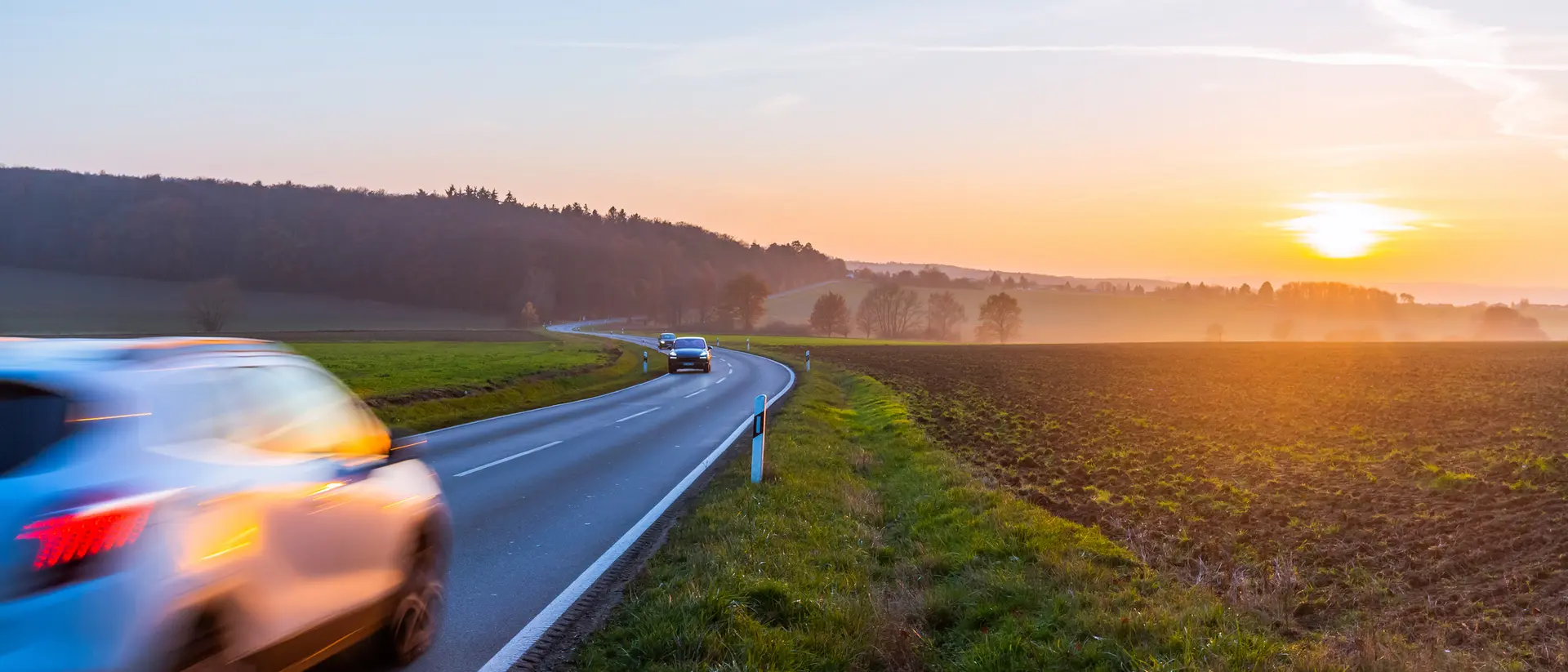 A car drives along a winding road at sunset, with fields on one side and trees in the background.