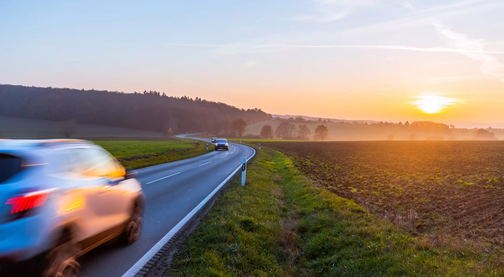 A car drives along a winding road at sunset, with fields on one side and trees in the background.