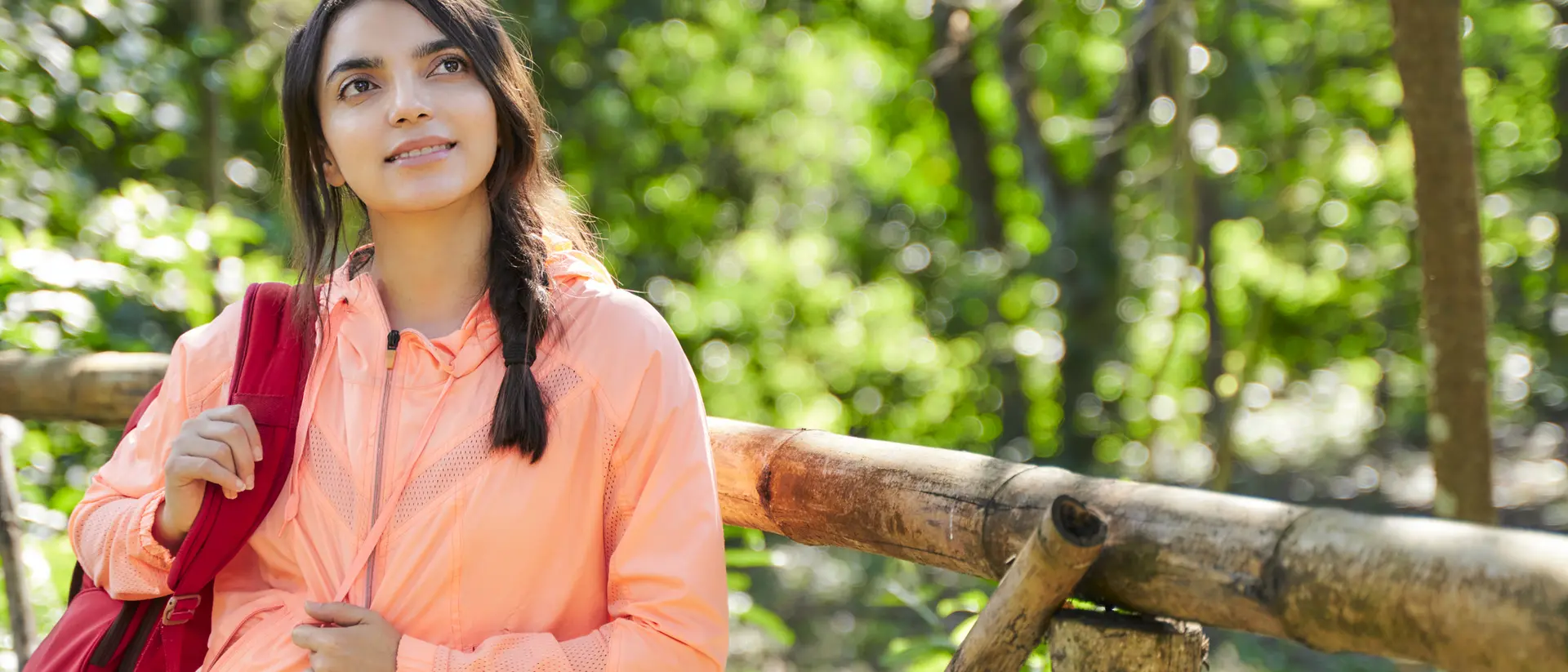 A young woman in an orange jacket stands by a wooden fence, looking thoughtfully into the distance in a lush forest.