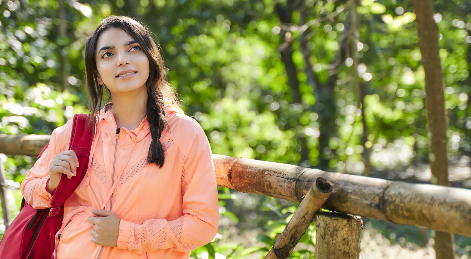 A young woman in an orange jacket stands by a wooden fence, looking thoughtfully into the distance in a lush forest.