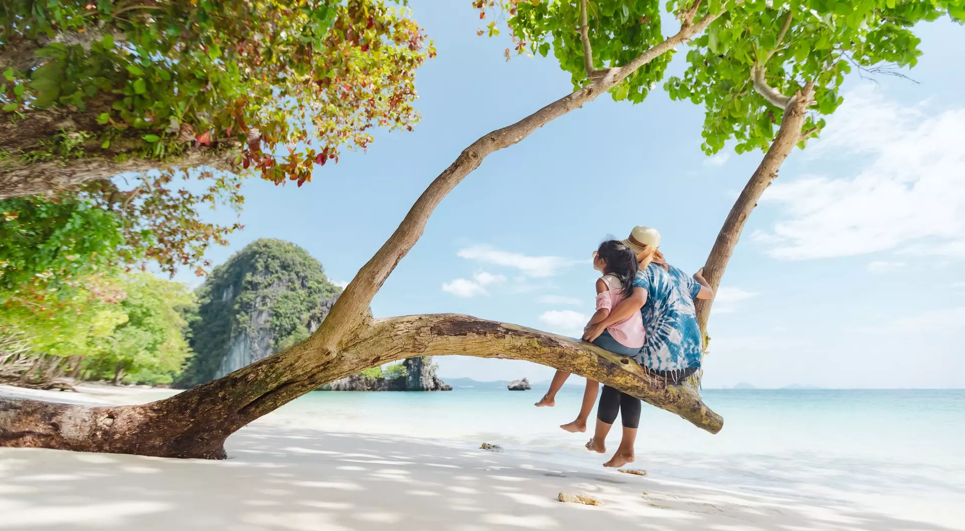 A woman sits on a tree branch by the beach, holding a child, with clear water and lush greenery in the background.